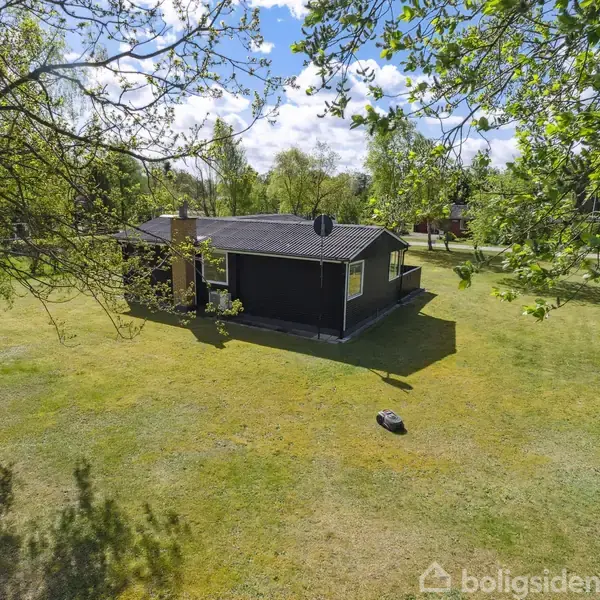 Et sort sommerhus står omgivet af en stor, grøn græsplæne med spredte træer. Solen skinner, og himlen er let skyet, hvilket giver en fredfyldt atmosfære.