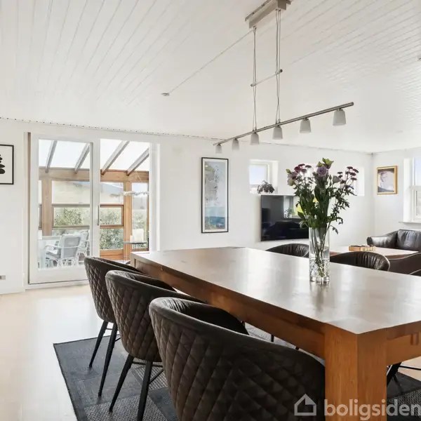 Spisebord med otte stole er placeret i en lys stue. Bordet har en vase med blomster, og der er abstrakte malerier på væggene. En åben veranda er synlig i baggrunden.
