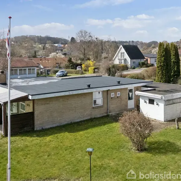 En enkeltstående bungalow med fladt tag og store vinduer, omgivet af en velplejet græsplæne og flagstang, beliggende i et skovklædt boligområde med bakker i baggrunden.