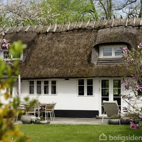 Hus med stråtag og hvide vægge, omgivet af en frodig have med blomstrende træer. Foran huset står et bord med stole. Baggrunden har grønne træer, der skaber en idyllisk stemning.