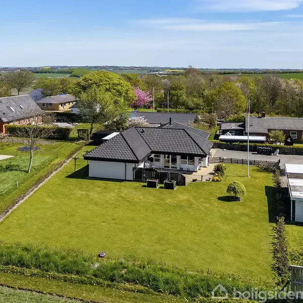 Et sort-hvidt enfamilieshus sidder stille midt i en stor grøn græsplæne. Omkranset af andre huse og marker, giver omgivelserne en landlig følelse.