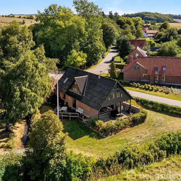 Rødt hus med stråtag står blandt træer og grønne overdækkede marker; en flagstang ses ved siden af huset. En stille vej snor sig gennem landskabet med flere huse i baggrunden.