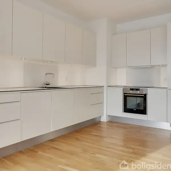 Hvidt, moderne køkken med indbygningsovn og underskabe. Køkkenet er velholdt og lyst med trægulv og LED-spots.