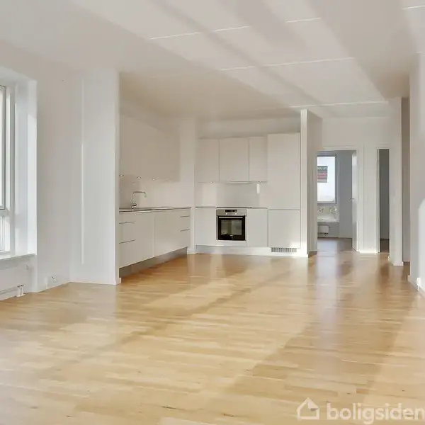 Stue med hvidt, åbent køkken. Trægulv dækker rummet. Store vinduer til venstre lader lys ind. En dør i baggrunden fører til et andet rum. Ingen tekst.