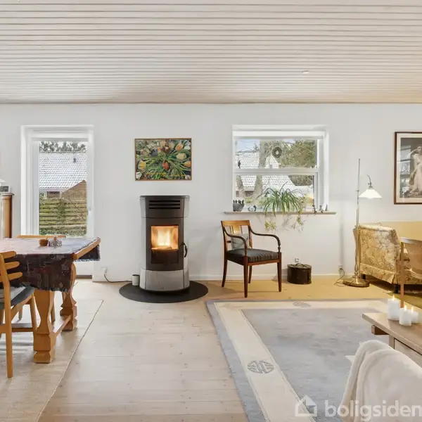 Et spise- og opholdsområde med brændeovn i midten, omgivet af et bord med stole, sofa, og kunst på væggene. Store vinduer giver naturligt lysindfald.