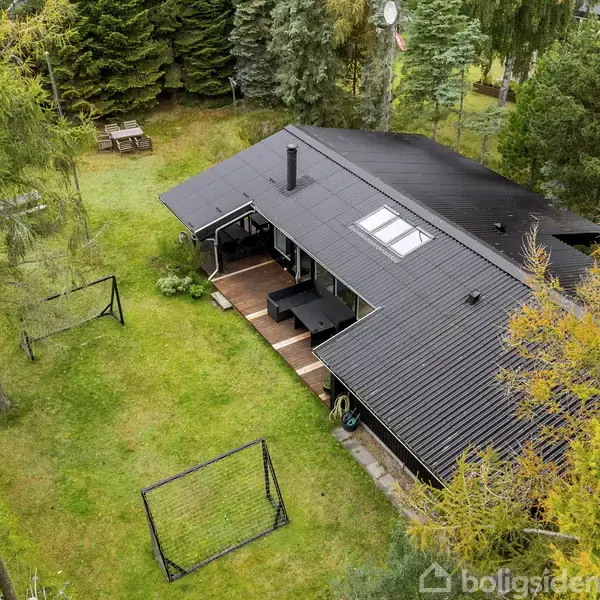 Hus med sort tag og terrasse har to fodboldmål i haven. Skovklædt område med træer og græs, samt picnicbord i baggrunden.