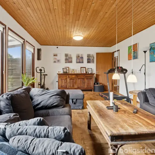 Stue med sorte sofaer centreret omkring et træsofabord; store vinduer til venstre lukker lys ind. Vægdekorationer og en trækommode ses i baggrunden. Lysebrunt loft og mørkt gulv.