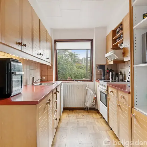 Køkken med brune træskabe og røde bordplader. En ovn står på bordpladen, og der er flere køkkenapparater. Vindue viser træer udenfor, og hylder indeholder mikrobølgeovn og flasker.