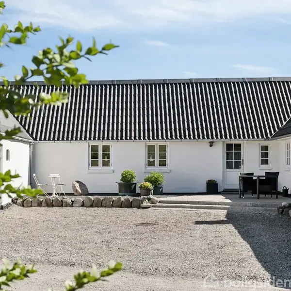 Hvidt hus med sort tag står i en gårdsplads med grus. Omkring er der grønne planter og en terrasse med havemøbler. Række af små buske langs fundamentet.