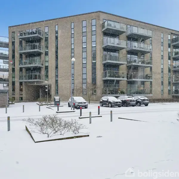 Bygning med flere etager og altaner står i et sneklædt område. Flere biler er parkeret foran bygningen. Sneen dækker jorden og omgivelserne med buske og lysstænger.