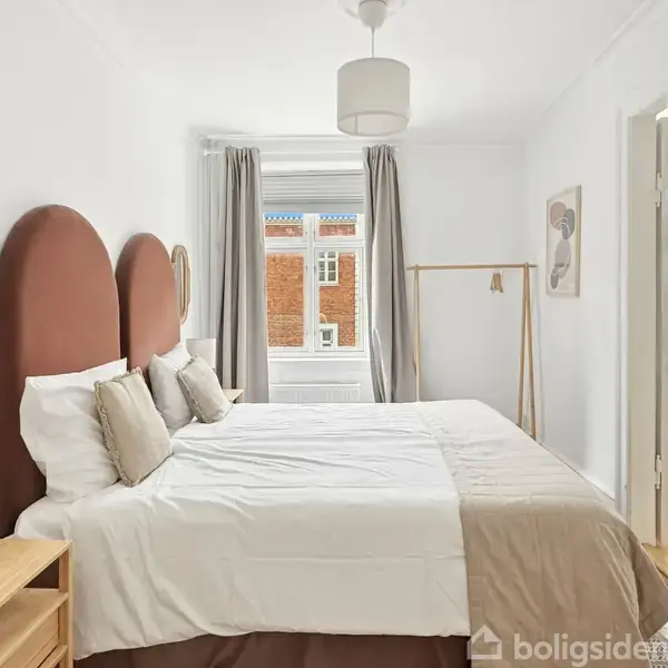 Soveværelse med hvide skabe åbner til en stue via franske døre, hvor der står et spisebord og stole. En kommode med planter står til højre. enkel indretning.