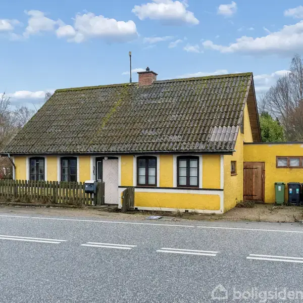 Gul, en-etagers hus med mørkt tag står ved vejkant. Trægitterhegn foran, omgivet af træer og buske. Fyldte affaldsspande ved siden af. Klar himmel med få skyer.