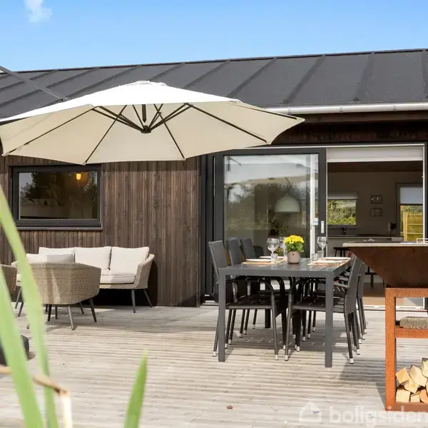 Terrasse med træbord og stole, solparasol og udendørs pejs med træopbevaring. Sofaarrangement ved huset. Græsset vokser langs kanten. Klar blå himmel i baggrunden.
