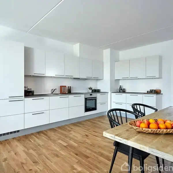 Et moderne køkken med hvide skabe og en indbygget ovn. En træskål med appelsiner står på et træbord. Der er en flettet plantekurv ved en blå væg.