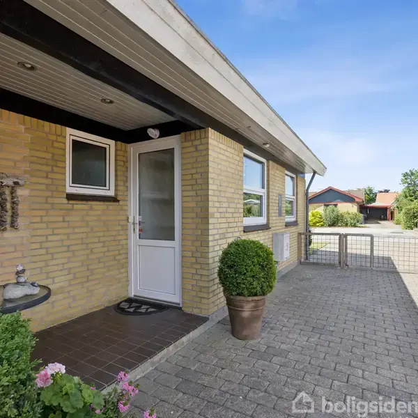 Hus i gule mursten med en hvid dør og store vinduer, placeret i en indkørsel med fliser. En krukke med grønt står ved døren. Omkringliggende huse og grønne træer i baggrunden.