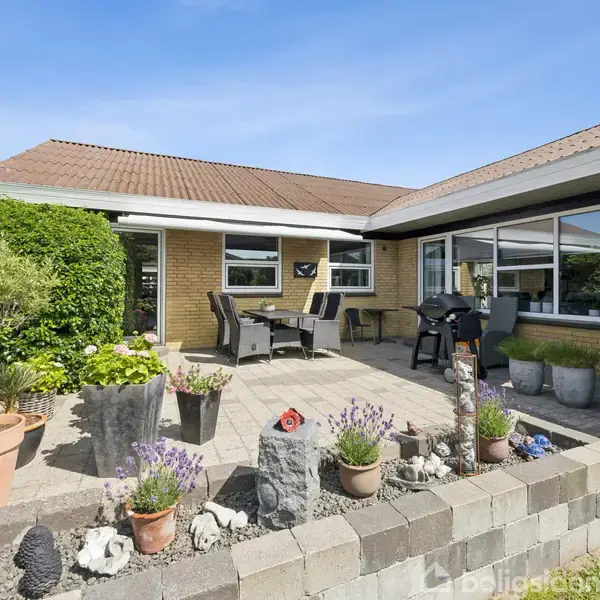 Terrasse med bord og stole er omgivet af planter og dekorationer. Haven er frodig med buske og blomster. Huset i baggrunden har store vinduer og brune murstensvægge.
