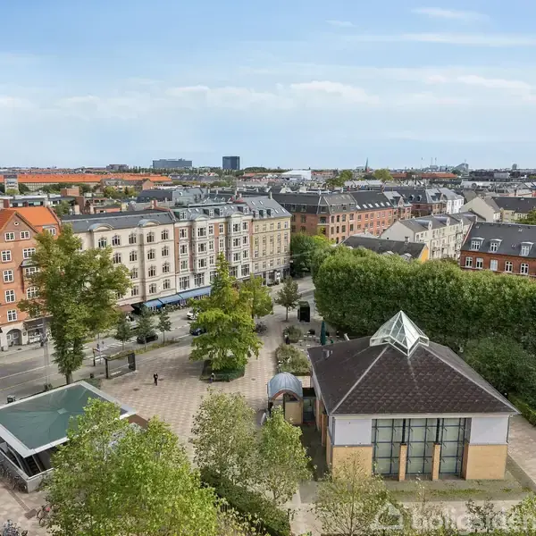 Bygning overser en byplads, omkranset af træer og flere ældre bygninger med røde tage. Konteksten viser en byudsigt under en blå himmel med spredte skyer.