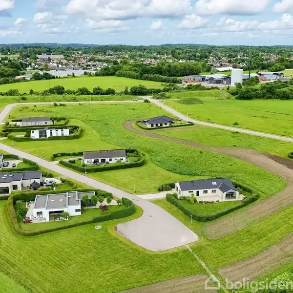 Flere moderne enfamiliehuse ligger spredt ud i et grønt område med velplejede haver. En vej snor sig mellem husene. I baggrunden ses det omkringliggende landskab og en by.