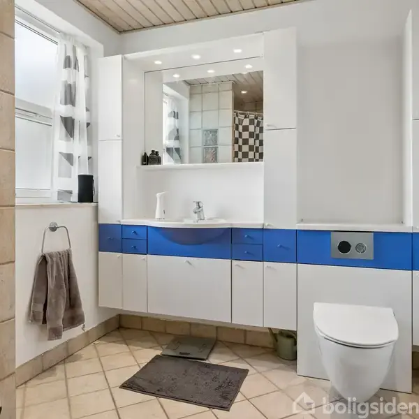 Badeværelse med hvidt vaskeskab, blå skuffer, og spejl med belysning. Toilet ved siden af. Hvide vægge, klinkegulv, håndklæde på holder, og vindue til venstre.