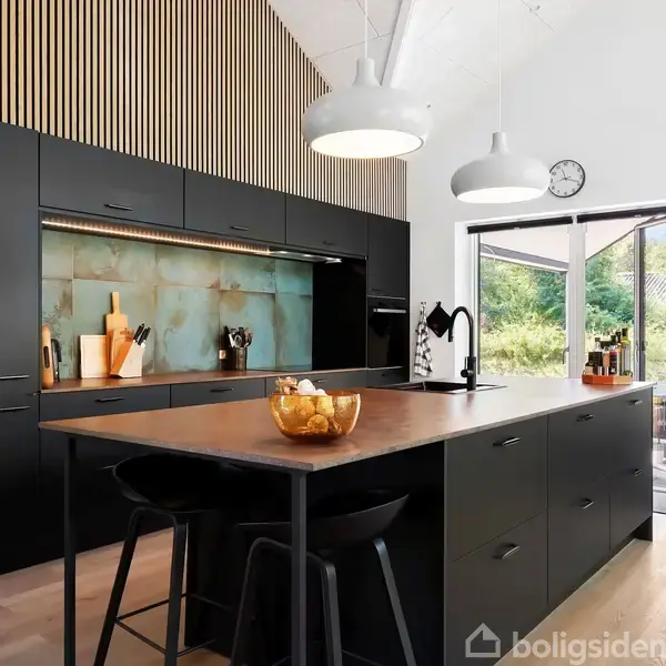 Moderne sort køkken tilfører elegance, med en stor køkkenø og indbyggede apparater. Rummet er oplyst af pendellamper, med store vinduer der vender ud mod en grøn have. En skål står på bordet.