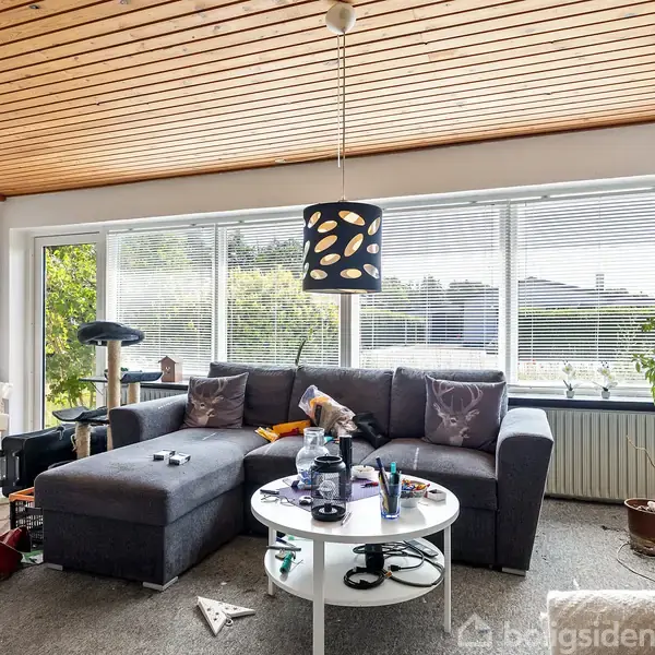 En grå hjørnesofa står i en stue. En rund sofabord foran er rodet. Store vinduer med persienner giver udsigt til en have. Plakater og plantere dekorerer rummet.