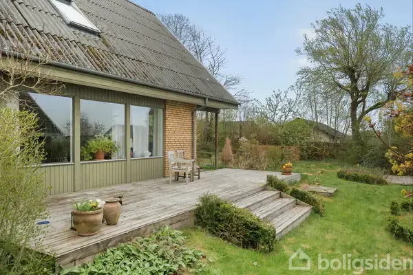 Et hus med en skråt tag og store vinduer har en træterrasse med to krukker og en stol. Haven er omgivet af buske og træer på en græsplæne.