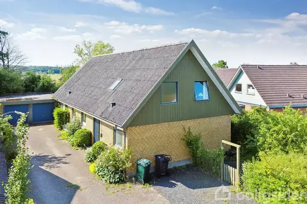 Et to-etagers hus med skråt tag står omgivet af grønne buske i et beboelseskvarter. To affaldsspande ses ved siden af en flisebelagt indkørsel, der fører til en dobbeltgarage.