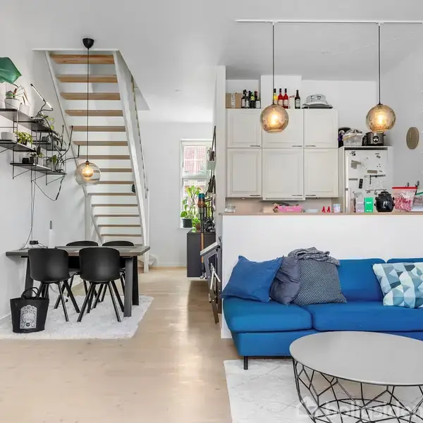 En stue med en blå hjørnesofa er placeret ved siden af et lavt, rundt sofabord. I baggrunden er et spisebord med sorte stole og en trappe til øverste etage.