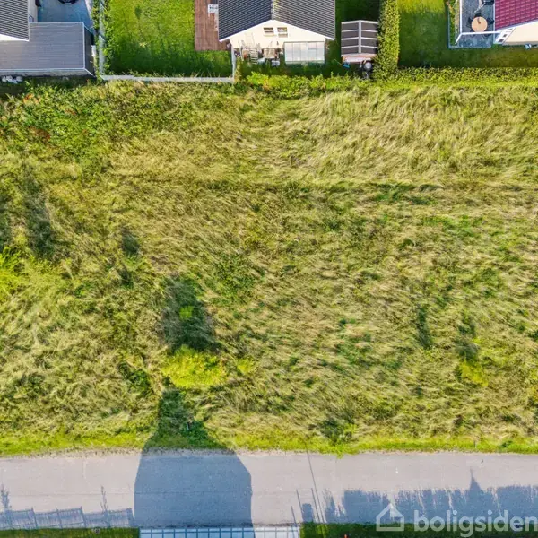 Græsareal set oppefra; græsset står højt og uplejet. Omkranset af huse på den ene side og en vej på den anden. Skygger fra træer og strukturer kaster på græsset.
