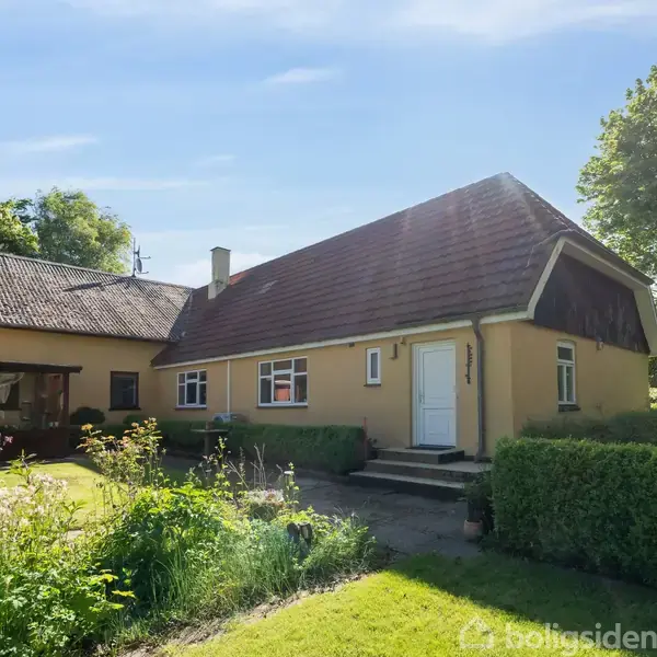 Hus står i solrigt haveområde med grønne træer og buske. Husets facade er gul med rødt tegltag, og en hvid dør er synlig på forsiden.