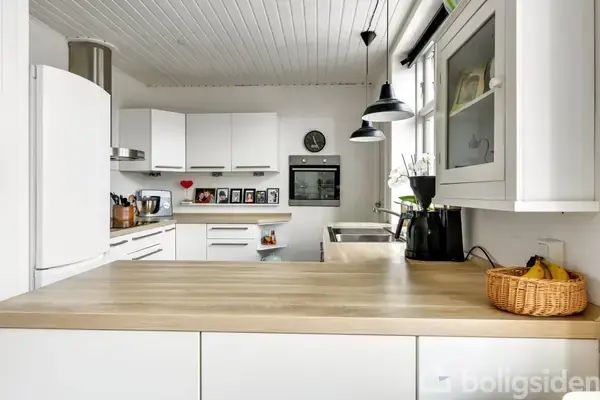 Et moderne køkken med træbordplade, hvide skabe og indbygningsovn. Der er en kurv med bananer samt blomster i en vase. Lamper hænger fra loftet. Fotos og ur ses.