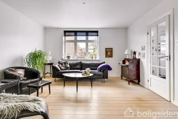 En stue med en sort sofa og et rundt sofabord i midten. Omkring står lænestole og en kommode. Store vinduer slipper lys ind, omgivet af lyse vægge og trægulv.