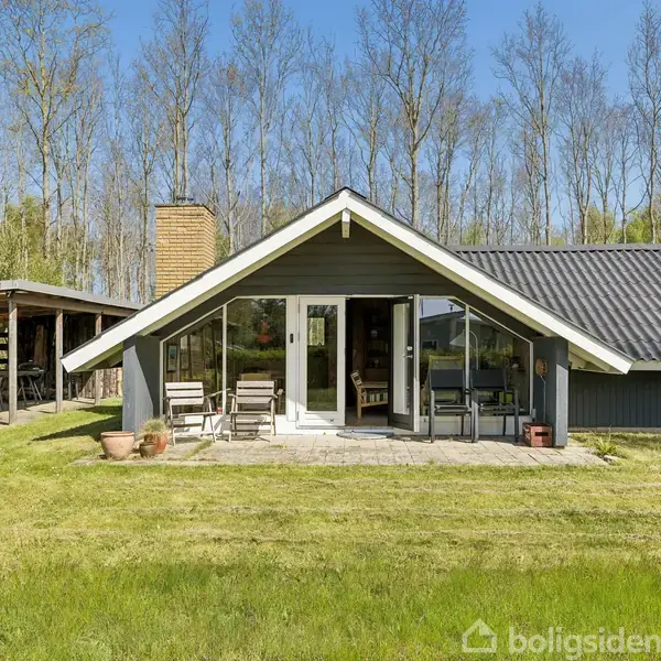 Sommerhus med mørkt tag og store vinduer står på en græsplæne. Havemøbler er placeret på terrassen. Der er en carport til venstre og træer i baggrunden. Ingen tekst i billedet.
