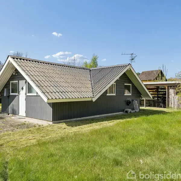 Et lille, gråt sommerhus med spidst tag står i en grøn have. Huset har flere små vinduer og er omgivet af træer og buske under en klar blå himmel.