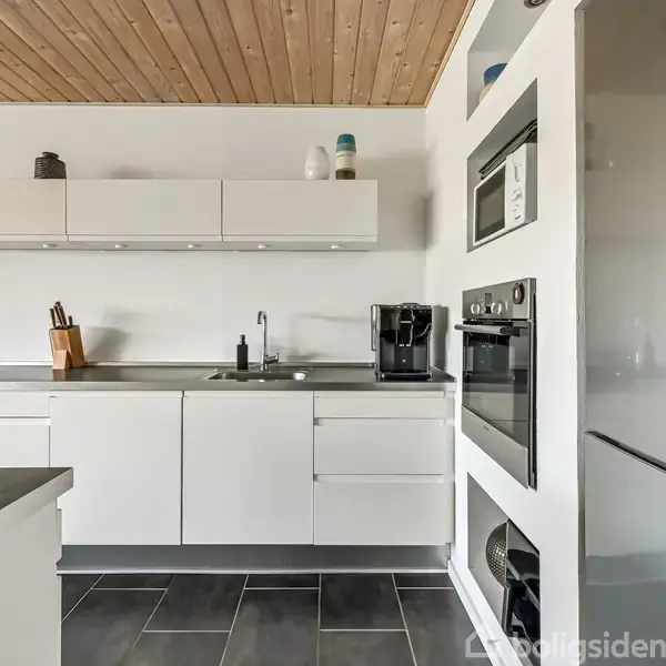 Et moderne køkken med hvide skabe og sorte bordplader. En indbygget ovn og mikrobølgeovn er synlige i højre side. Loftet er af træ, og gulvet er flisebelagt.