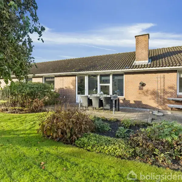 Rødt murstenshus med fladt tag har en terrasse foran med havemøbler. Det omkranses af en velplejet have med grøn græsplæne og buske, i en rolig, landlig omgivelser under blå himmel.