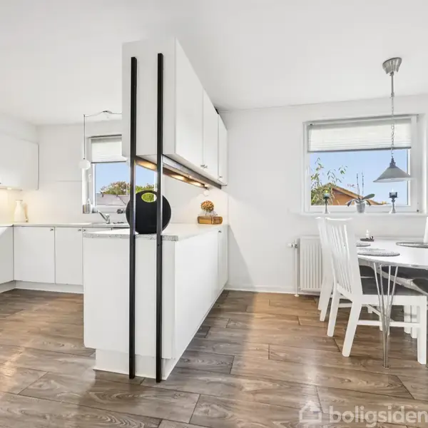 Køkken med hvide skabe står i centrum, flankeret af et spisebord og hvide stole ved siden af et vindue. Trægulve og naturligt lys præger det lyse rum.