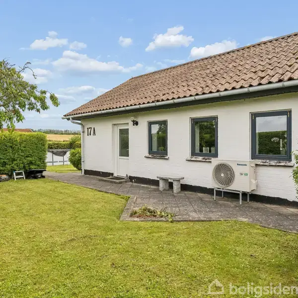 Huset har hvide vægge og et rødt tegltag. Det står på en velplejet græsplæne med flisebelagt terrasse. Dørnummeret "17A" er synligt. Træer og buske omkranser området.