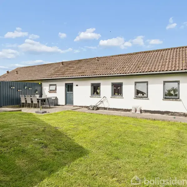 Et hvidt hus med brunt tegltag står i en have med grøn græsplæne. Der er et tørrestativ og et udendørs spisebord nær en mørk plankevæg.