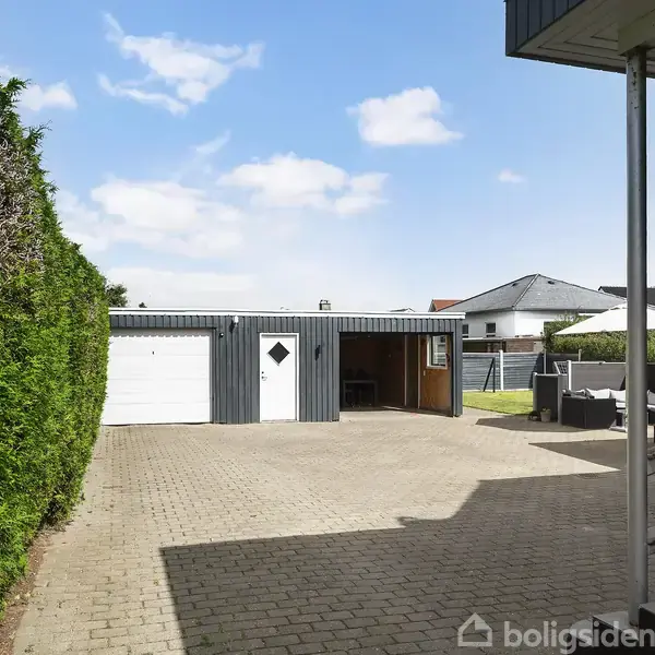 Garagebygning står for enden af en brostensbelagt indkørsel. En hæk til venstre og husvæg til højre omgiver området. Himlen er blå med skyer.