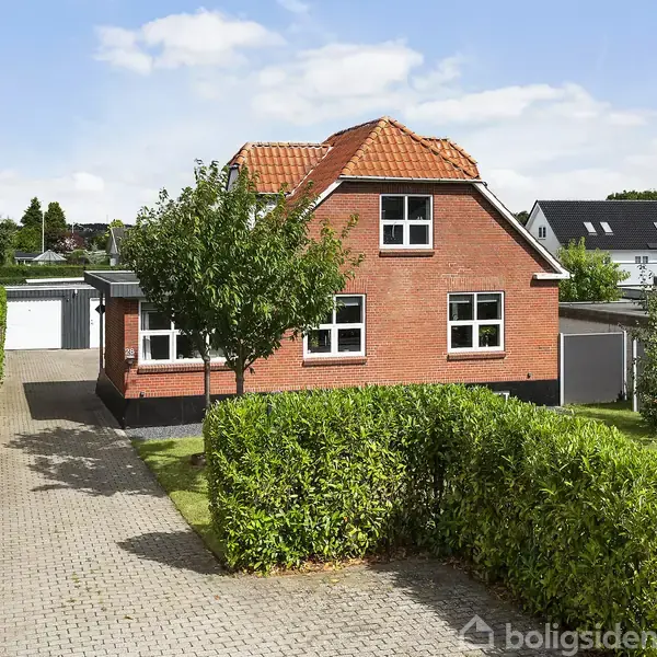 Rødt murstenshus med rødt tegltag står forrest. En lille træ vokser foran huset. Flisebelagt indkørsel og hæk omkranser haven, med garager synlige i baggrunden.
