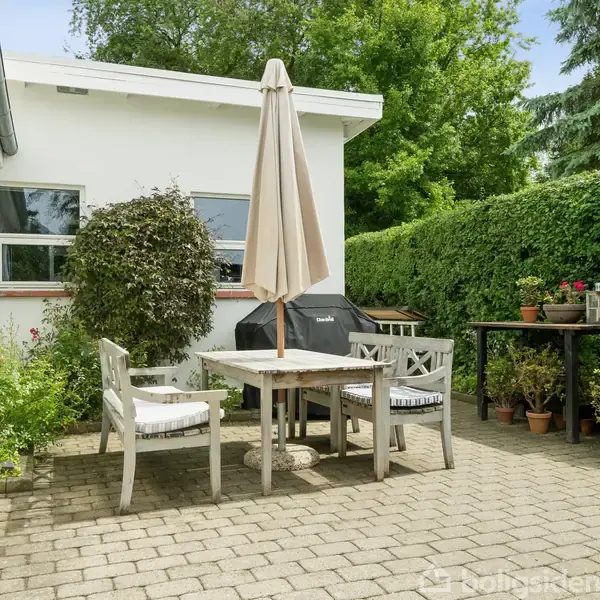 Terrasse med bord og stole i en gårdhave omgivet af grønne planter og buske. En stor parasol står midt på bordet, i nærheden af en sort grill.
