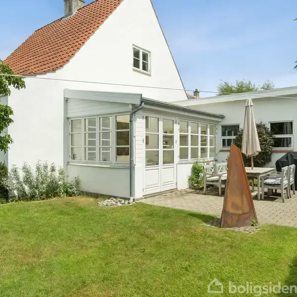 Hus med hvid facade og rødt tag ved en grøn græsplæne. Terrasse med havemøbler under en parasol. Omkranset af træer og buske.