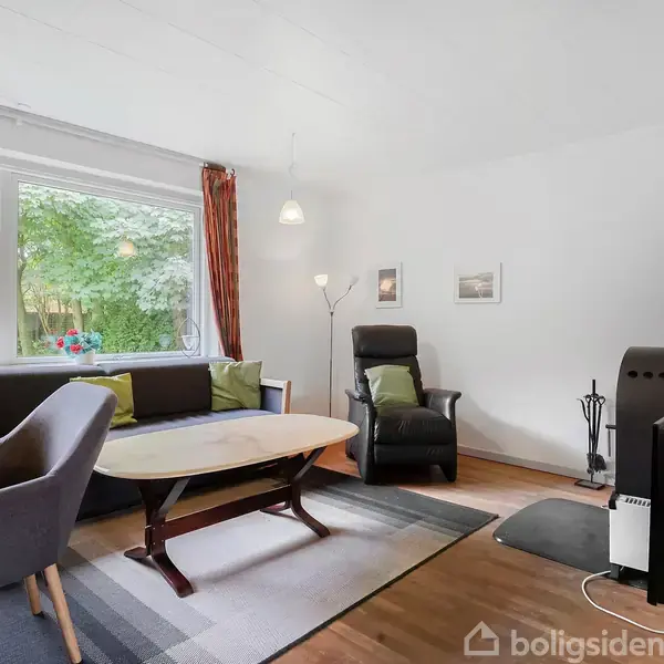 En stue har en sort lænestol, sofa med puder, sofabord, og et brændeovnshjørne. Store vinduer med udsigt til grønt. Trægulv og vægdekorationer skaber en hyggelig atmosfære.