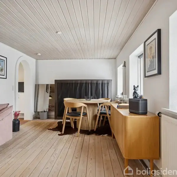 Et spisebord med stole står i et lyst rum med trægulv. En reol med pynt og et vindue med udsigt omkranser området. En sofa ses i baggrunden.