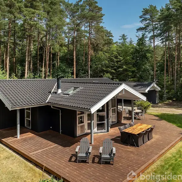 Sommerhus med træterrasse omgivet af skov; to stole og et bord står på terrassen. Skov trækker sig tæt rundt om huset, der har en mørk facade og gråtag.