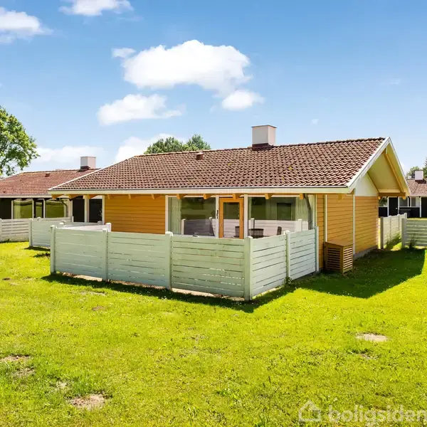 En gulmalet bungalow med en brun flisetag står omkranset af en hvid træhegn. Den ligger på en stor, grøn græsplæne under en klar blå himmel med spredte skyer.