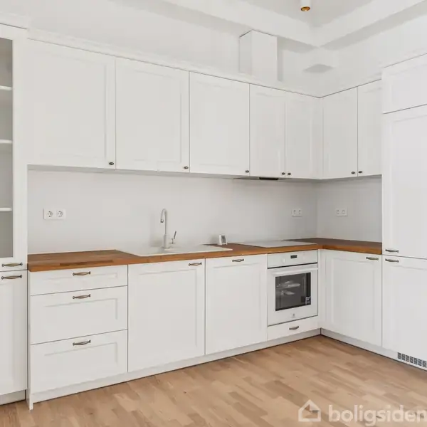 Et moderne hvidt køkken med træbordplade, indeholder indbygget ovn, vask, og overskabe. Lys trægulv og hvide vægge skaber et rent og minimalistisk miljø.