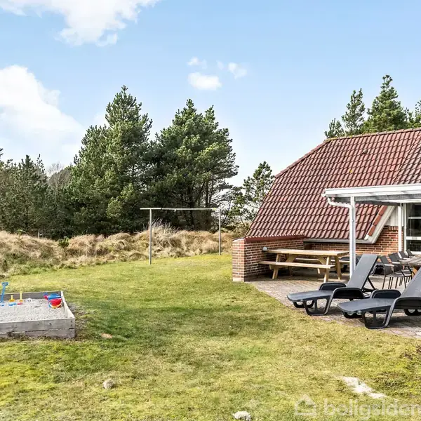 Hus med rød tagsten, terrasse med liggestole og havebord. Græsplæne med sandkasse og legetøj. Omgivet af skov og blå himmel.