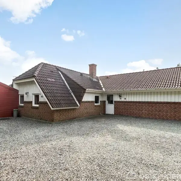 Et rødt murstenshus med tegltag står foran en grusbelagt indkørsel. Omgivet af træer og åben himmel, er der også en lille rød tilbygning til venstre.